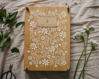 May include: A wooden sign with a floral design and the text "BLUE BRONTIDE FLOWERS". The sign is light brown with white floral patterns and is secured with metal fasteners. Flowers and a pair of scissors are arranged around the sign.