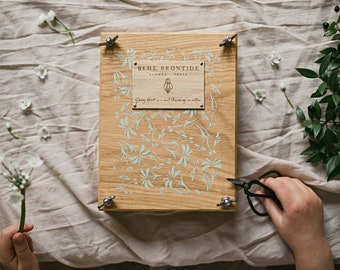 May include: A wooden flower press with the text "BLUE BRONTIDE FLOWER PRESS" and a floral design. The press is secured with metal hardware. A hand holds a sprig of white flowers, and another hand holds black scissors. The press is on a beige linen surface.