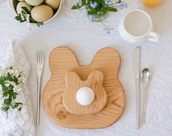 May include: A wooden bunny-shaped egg holder with a white egg, a bowl of eggs, a glass of orange juice, and a white mug on a white tablecloth. A fork, knife, and spoon are set next to the egg holder. Flowers and a napkin complete the Easter table setting.