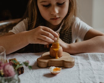 May include: A wooden egg holder shaped like a car, holding a soft-boiled egg. A child is dipping a piece of toast into the egg. The table setting includes a glass of juice, flowers, and bread.