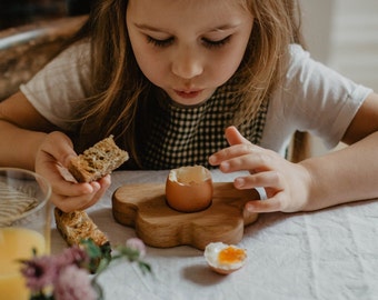 May include: A wooden egg holder shaped like a flower holds a brown eggshell. A slice of bread is held in a hand. A half-eaten egg with a yolk is on the table. The scene is set on a white tablecloth.