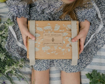 May include: A wooden box with a floral design and the text "BLUE BRONTIDE". The box is held by a person wearing a blue floral dress. The box has light blue floral patterns and beige straps.