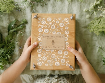 May include: Wooden flower press with white daisy pattern and metal hardware. The press has a rectangular wooden plaque with the text "BLUE BRONTIDE FLOWER PRESS" and a quote. The press is surrounded by greenery.