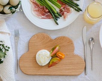 May include: A wooden cloud-shaped serving board holds a ball of mozzarella cheese and sliced tomatoes. The table setting includes plates of asparagus and prosciutto, a jar of liquid, and a bowl of eggs. A white napkin with a floral arrangement is on the left.