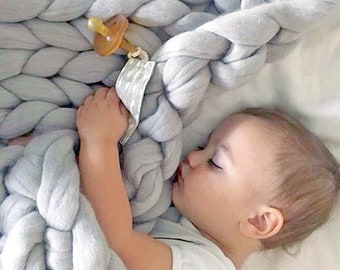 May include: A baby sleeping next to a large, chunky knit blanket. The blanket is a light gray color and has a braided design. A pacifier is attached to the blanket. The baby is wearing a white onesie.