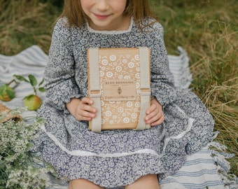 May include: A young person holding a wooden box with a daisy pattern and the text "BLUE FRONTIDE". The box has beige straps and is surrounded by grass and a blue floral dress. The box appears to be a picnic basket.