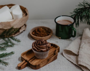 May include: A chocolate cupcake with chocolate frosting sits on a wooden serving board. A green enamel mug of hot chocolate, a small bowl of cocoa powder, and a bag of marshmallows are also on the table. The scene is decorated with pine branches and a linen napkin.