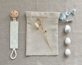 May include: A wooden baby pacifier clip with a light blue and beige patterned strap, a beige drawstring bag, and three speckled decorative eggs. A sprig of dried flowers rests on the bag. The items are arranged on a gray linen surface.