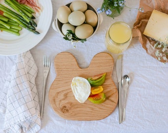 May include: A wooden bunny-shaped cutting board holds burrata cheese and sliced tomatoes. A plate of asparagus and prosciutto, a bowl of eggs, and a jar of juice are on a white tablecloth. Silverware and a checkered napkin complete the scene.