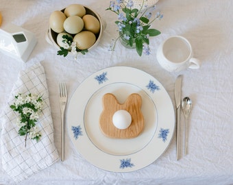 May include: A festive Easter table setting with a white tablecloth. A plate with blue floral accents holds a wooden bunny-shaped egg holder with a white egg. Silverware, a napkin with flowers, and a bowl of eggs complete the scene.