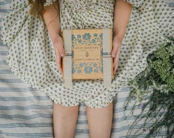 May include: A wooden box with blue floral designs and the text "BLUE BRONTIDE FLOWER SOAPS" is held by a person. The box is secured with beige straps and metal clasps. The person is wearing a white dress with a floral pattern.