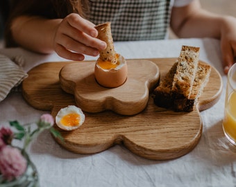 May include: A wooden breakfast tray with a flower-like design holds a soft-boiled egg, toast, and a half-cut egg. The tray is made of light-colored wood. A person is dipping toast into the egg. A glass of orange juice is visible.