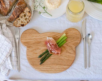 May include: A wooden bear-shaped serving board with asparagus and prosciutto. The board is surrounded by a fork, knife, spoon, bread, cheese, and a glass jar of liquid. The items are arranged on a white tablecloth.
