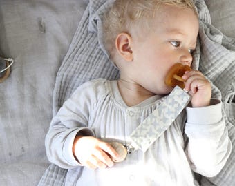 May include: A baby wearing a light gray onesie and sucking on a natural rubber pacifier. The pacifier has a butterfly-patterned strap. The baby is lying on a gray blanket.