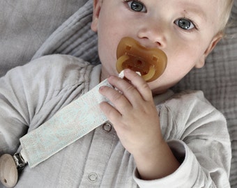 May include: A baby wearing a light gray onesie with a pacifier in its mouth. The pacifier is attached to a light blue and white patterned strap with a wooden clip.