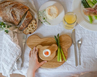 May include: Overhead shot of a breakfast table set with a loaf of sliced bread, a plate of asparagus and prosciutto, and a glass of juice. A wooden bear-shaped serving board holds a small dish with an egg. A child's hand is reaching for the board.