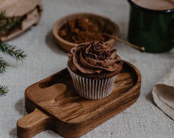 May include: A chocolate cupcake with chocolate frosting and sprinkles sits on a wooden serving board. A cup of hot chocolate and cocoa powder are in the background. The cupcake is in a white paper liner.