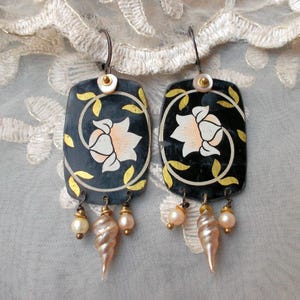 Può includere: Un paio di orecchini fatti a mano. Ogni orecchino presenta un ciondolo rettangolare blu scuro con un motivo floreale bianco e pesca, accentuato da foglie color oro. Gli orecchini pendono con perle e un ciondolo a forma di conchiglia.