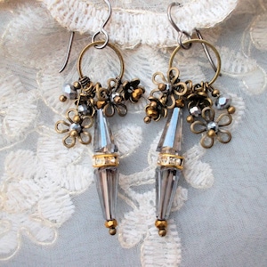 Könnte beinhalten: Ein Paar Ohrhänger im Vintage-Stil. Jeder Ohrring hat einen klaren, facettierten Kristallanhänger mit goldfarbenen Akzenten. Kleine, blütenförmige Charms und Perlen in Silber- und Goldtönen schmücken die Oberseite, befestigt an einem kreisförmigen Reifen.