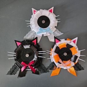 May include: Three felt cat head brooches. One is white with a blue bow, one is black with a pink bow, and one is orange and white with a pink bow.
