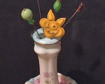 May include: A white porcelain vase with a floral design and a gold trim, holding a collection of vintage-style pins with colourful beads and a yellow flower-shaped pin.