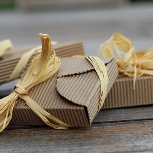 May include: Three small, brown cardboard boxes tied with natural raffia ribbon. The boxes are stacked on top of each other, with the top box slightly offset to the side. The boxes are all tied with the same type of ribbon, which is a natural, straw-like material.