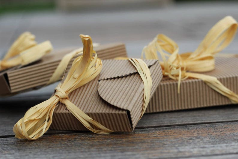 May include: Three small, brown cardboard boxes tied with natural raffia ribbon. The boxes are stacked on top of each other, with the top box slightly offset to the side. The boxes are all tied with the same type of ribbon, which is a natural, straw-like material.