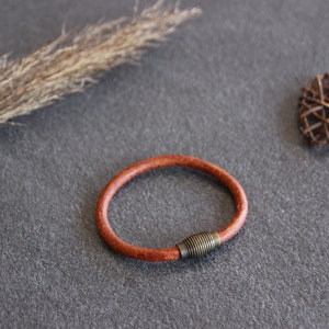 Puede incluir: Un brazalete de cuero marr&oacute;n con un cierre de metal. El brazalete est&aacute; sobre una superficie gris.