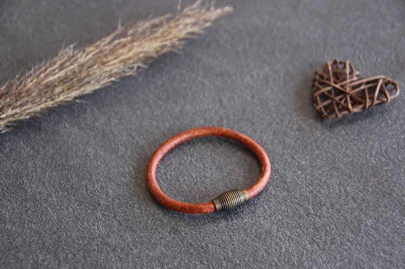 Puede incluir: Un brazalete de cuero marr&oacute;n con un cierre de metal. El brazalete est&aacute; sobre una superficie gris.