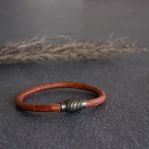 Puede incluir: Un brazalete de cuero marr&oacute;n con un cierre de metal plateado. El brazalete est&aacute; sobre una superficie gris.