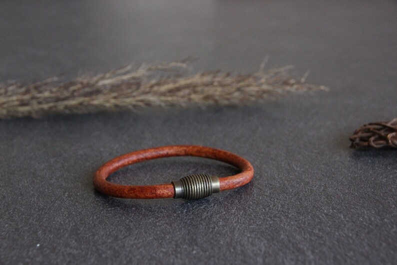 Puede incluir: Un brazalete de cuero marr&oacute;n con un cierre de metal plateado. El brazalete est&aacute; sobre una superficie gris.