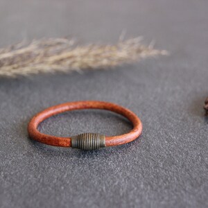 Puede incluir: Un brazalete de cuero marr&oacute;n con un acento met&aacute;lico. El brazalete est&aacute; sobre una superficie gris.