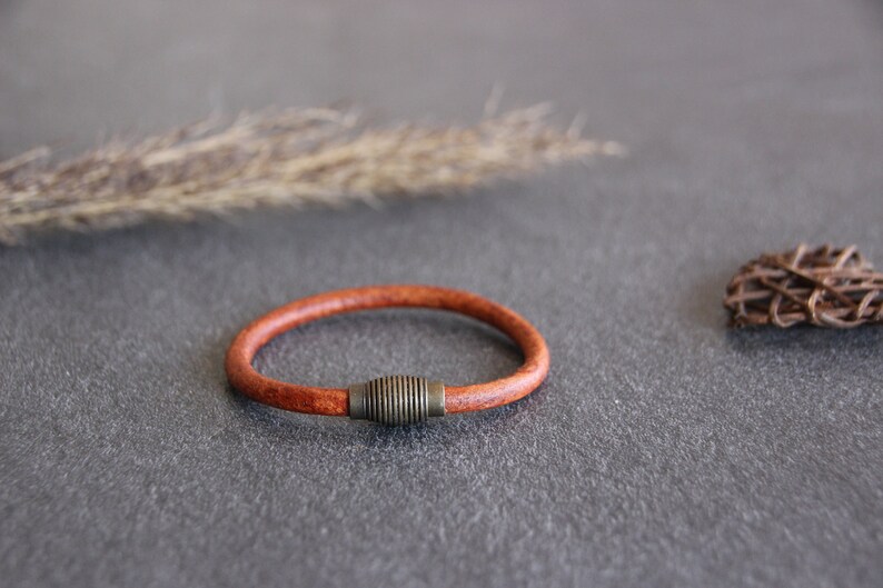 Puede incluir: Un brazalete de cuero marr&oacute;n con un acento met&aacute;lico. El brazalete est&aacute; sobre una superficie gris.