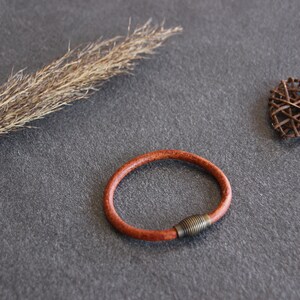 Puede incluir: Un brazalete de cuero marr&oacute;n con un cierre de metal. El brazalete est&aacute; sobre una superficie gris con un tallo de hierba seca y un coraz&oacute;n tejido.