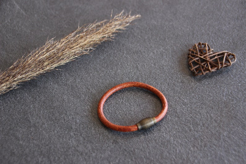 Puede incluir: Un brazalete de cuero marr&oacute;n con un cierre de metal. El brazalete est&aacute; sobre una superficie gris con un tallo de hierba seca y un coraz&oacute;n tejido.