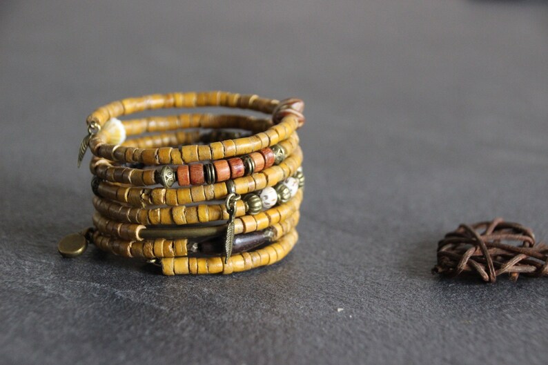 May include: A multi-strand bracelet made of yellow wood beads with brown and white accents. The bracelet has several metal charms, including a feather and a small disc.