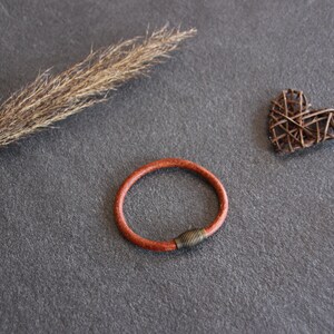 Puede incluir: Un brazalete de cuero marr&oacute;n con un cierre de metal. El brazalete est&aacute; sobre una superficie gris con un tallo de hierba seca y un coraz&oacute;n tejido.