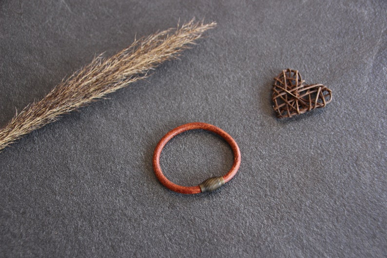 Puede incluir: Un brazalete de cuero marr&oacute;n con un cierre de metal. El brazalete est&aacute; sobre una superficie gris con un tallo de hierba seca y un coraz&oacute;n tejido.