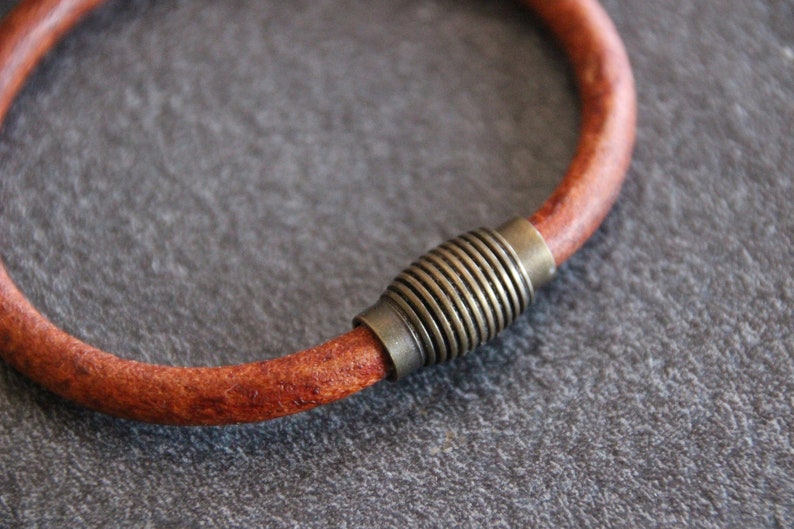 Puede incluir: Un brazalete de cuero marr&oacute;n con un cierre magn&eacute;tico de lat&oacute;n. El brazalete est&aacute; hecho de una sola pieza de cuero y tiene un dise&ntilde;o simple y minimalista.