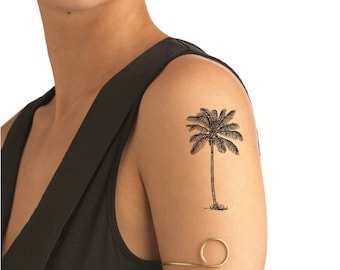 May include: A black and white palm tree tattoo on a person's upper arm. The person is wearing a black sleeveless shirt and a gold arm cuff.