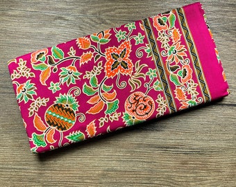 May include: Folded magenta fabric with a floral pattern in orange, green, and black. The design features stylized flowers and leaves. A magenta border runs along one edge. The fabric is likely cotton or a similar material.