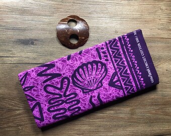 May include: A folded purple fabric with a seashell and tribal pattern, and a brown coconut shell ornament. The fabric has a dark purple design on a lighter purple background. The fabric's edge has text in a foreign language.