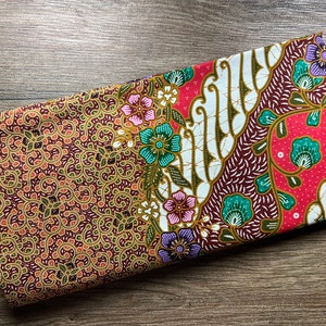 May include: Folded fabric with a detailed batik design. The fabric features a combination of floral and abstract patterns in shades of red, brown, white, green, and purple. The design is intricate and colourful, showcasing a traditional textile art form.