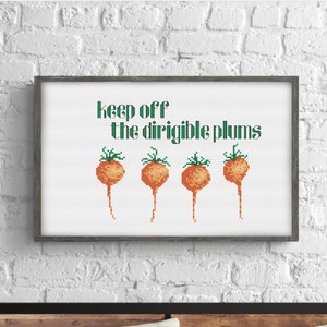 以下が含まれることがあります： 緑色の文字で「keep off the dirigible plums」と書かれた額縁付きのクロスステッチデザイン。オレンジと緑のプラムが4つ、テキストの下に刺繍されています。