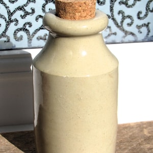 May include: A small, pale green ceramic jar with a cork stopper. The jar has a cylindrical body and a rounded top. The cork stopper is a light brown colour. The jar is on a wooden surface.