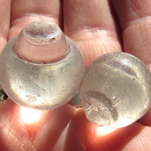 May include: Two clear glass bottle stoppers. One has a rounded top and a circular base. The other is a sphere. Both are held in a person's hand.