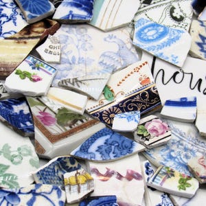 May include: A selection of broken ceramic fragments featuring floral and geometric designs in blue, white, pink, and green. Some pieces have gold edging. One fragment has the word "house" written on it.