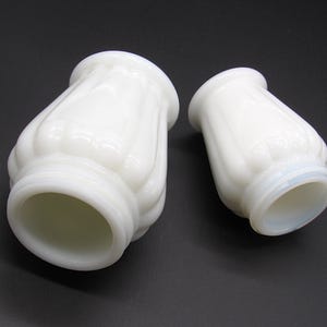 May include: Two white milk glass lamp shades with a fluted, vertical ribbed design. The shades feature a wide, open circular top and a slightly narrower base with a smooth, rounded rim, showcasing a translucent, milky appearance throughout.