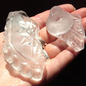 May include: Two clear, faceted glass fragments with intricate geometric patterns. The larger piece features a star-like design and a row of circular indentations, while the smaller piece has a smooth, rounded top and a textured, diamond-cut base.