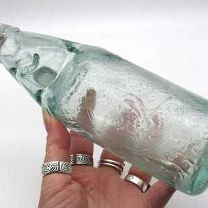 May include: A vintage light green glass bottle with a cork stopper. The bottle features embossed text and a circular emblem on the side. The glass has a slightly textured, bubbly appearance, characteristic of antique soda bottles from the early 20th century.